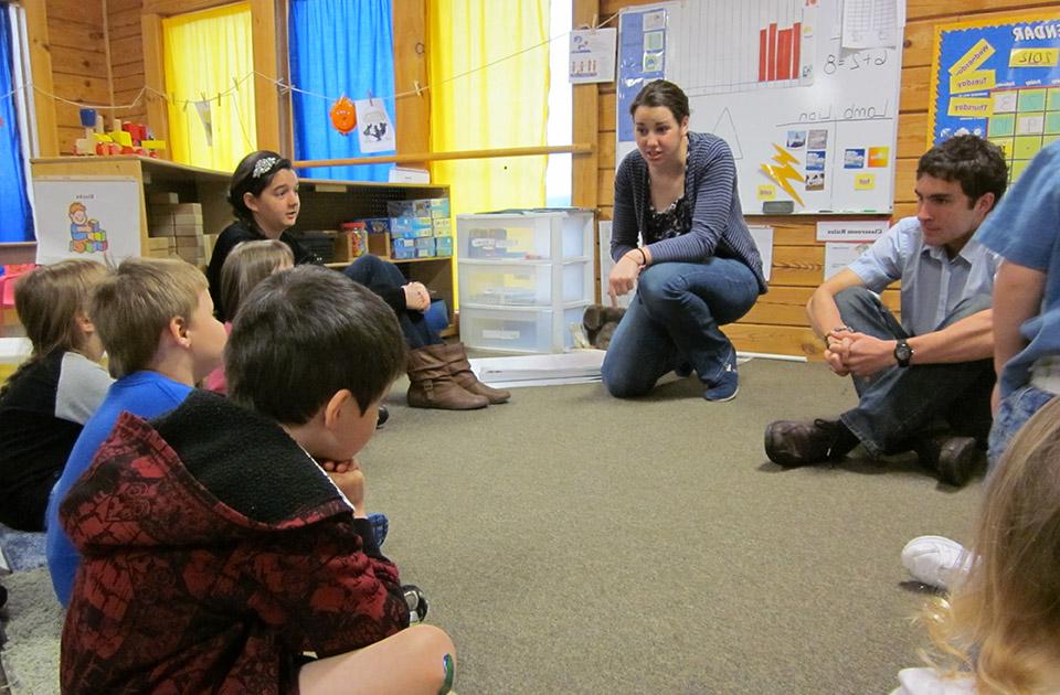 学生们在当地学校打工. Two 博彩网址大全 students sit on the floor at the front of a classroom with younger students gathered in a semi-circle.